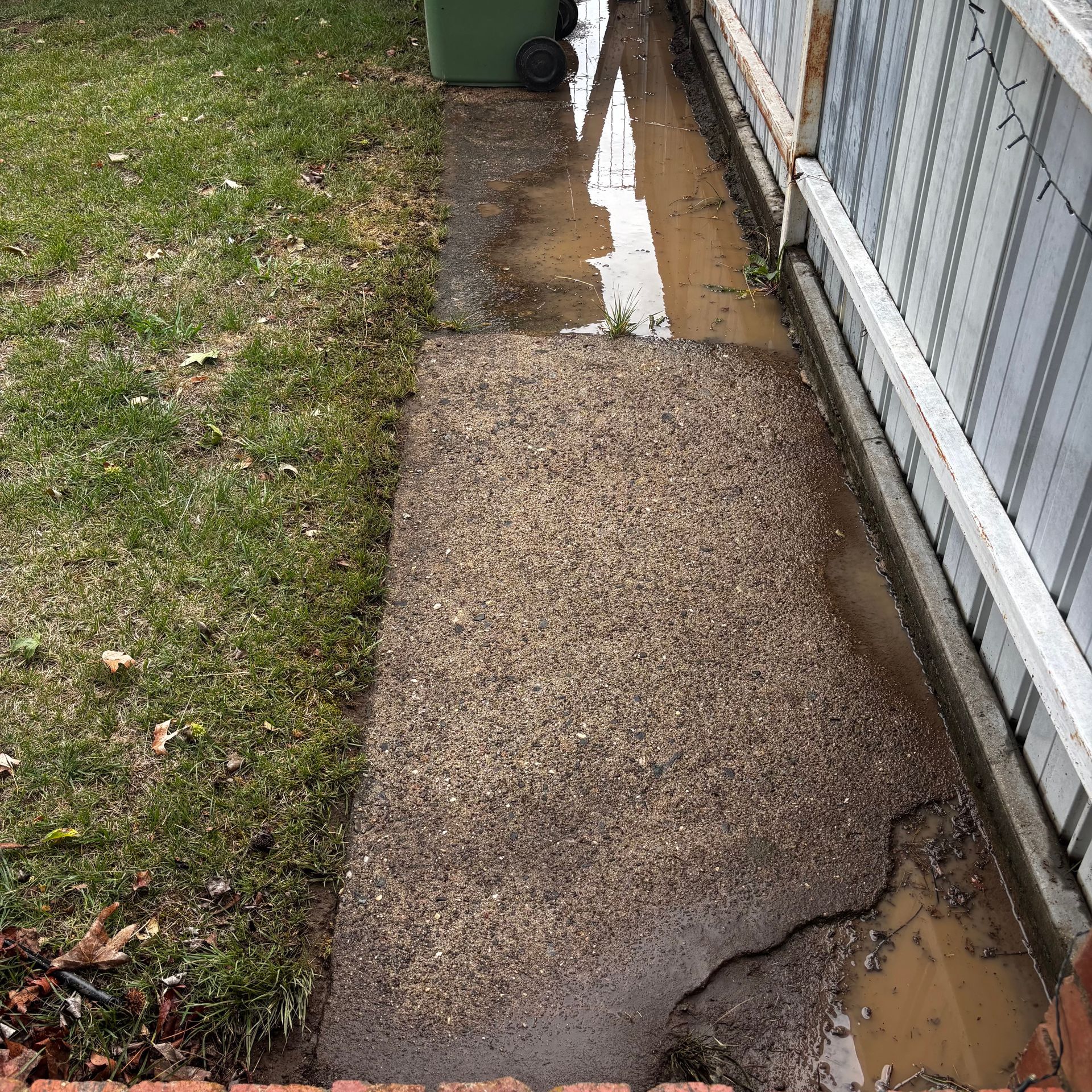 Damaged concrete walkway beside a fence, with standing water along the edge and grass on the left.