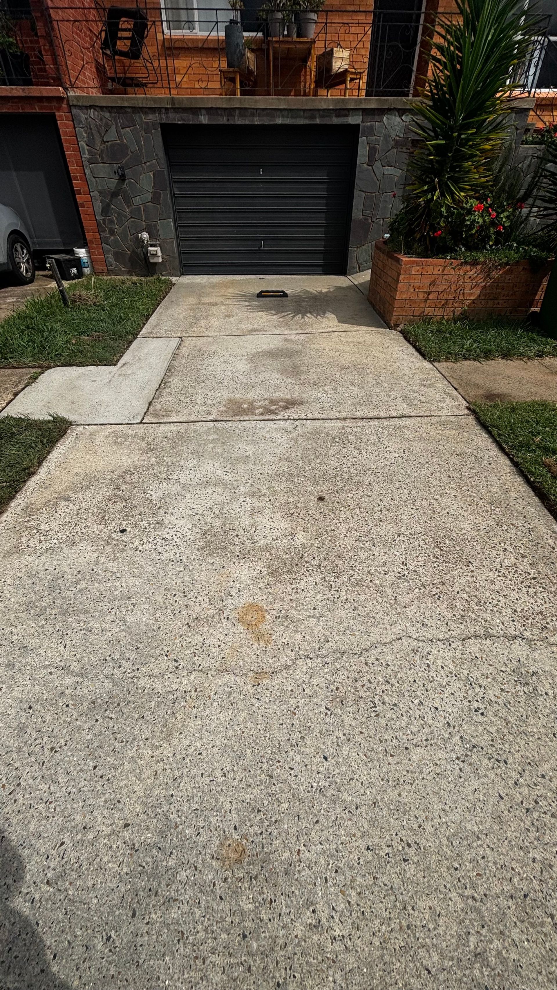Concrete driveway leading to a black garage door beside brick homes and small front gardens
