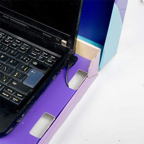 A black laptop sits inside a purple cardboard box, with a power cable connected to the side.