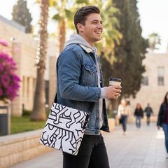 אדם מחייך בז'קט ג'ינס וקפוצ'ון אפור נושא תיק שליח עם דוגמאות בשחור-לבן, הולך בחוץ.