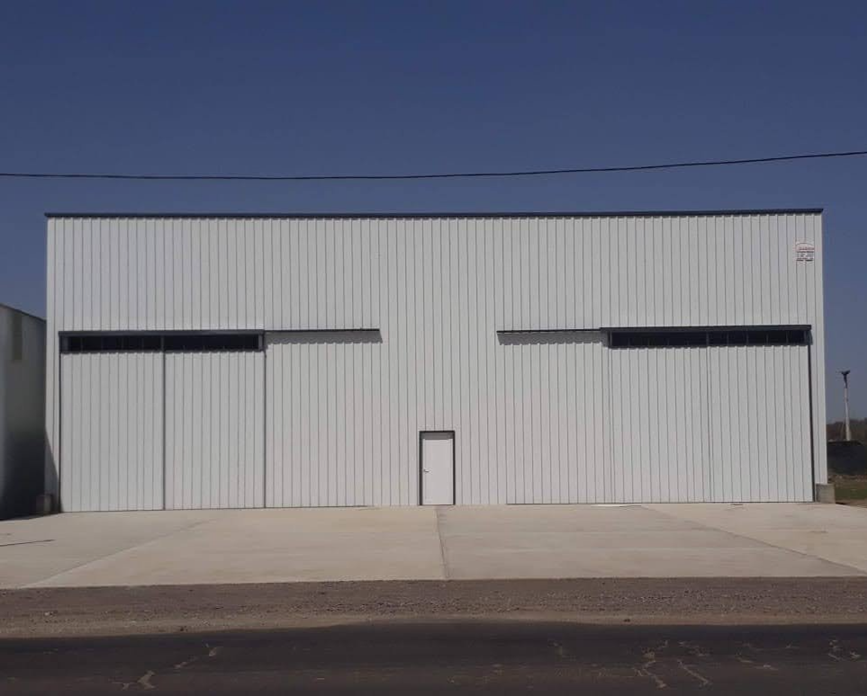 Nave industrial blanca con una pequeña puerta central y estrechas ventanas horizontales bajo un cielo despejado.