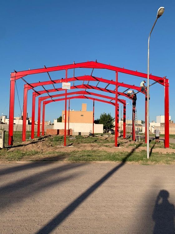 Estructura de acero rojo en construcción en un terreno cubierto de hierba junto a una carretera, bajo un cielo azul despejado.