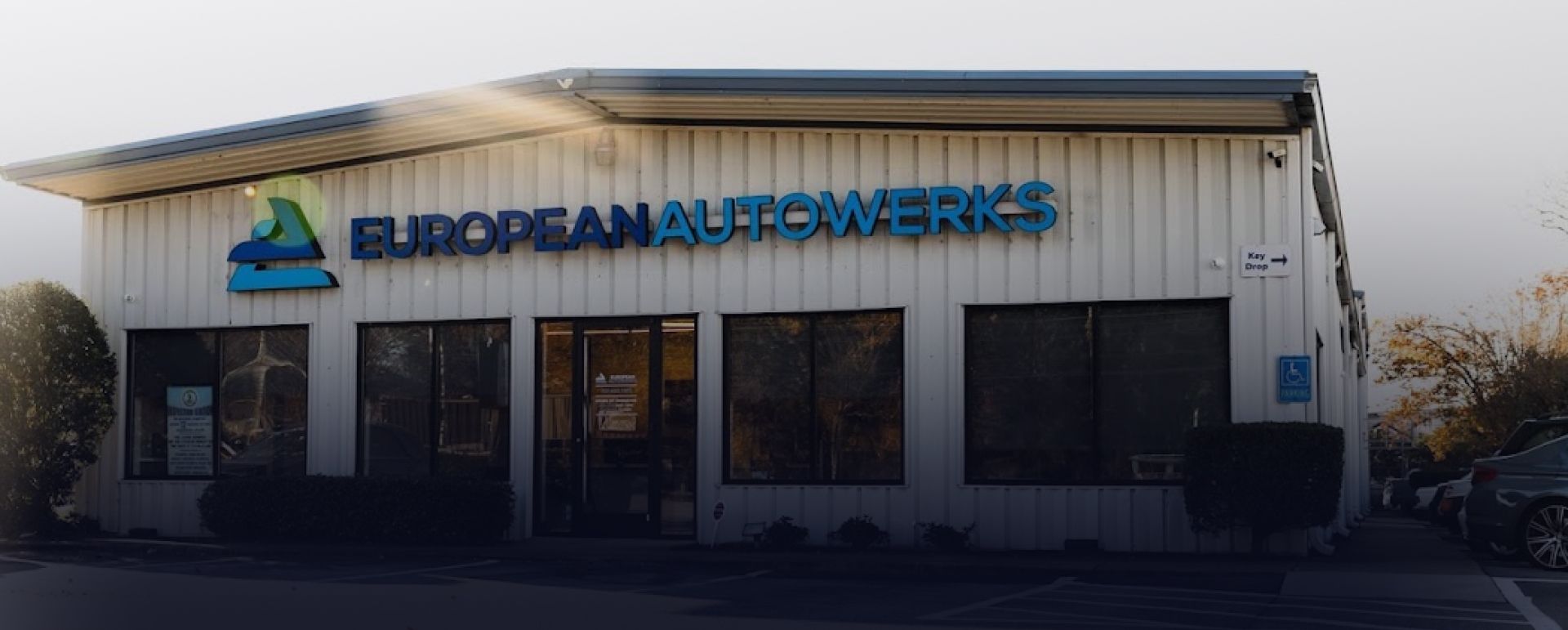 Exterior of European Auto Warehouse storefront with blue sign and glass windows at dusk | European Autowerks – Virginia Beach