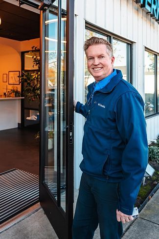 Smiling person in a blue jacket standing in a doorway outside a building | European Autowerks – Virginia Beach