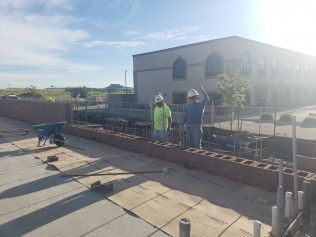 Two construction workers are working on a brick wall in front of a large building.