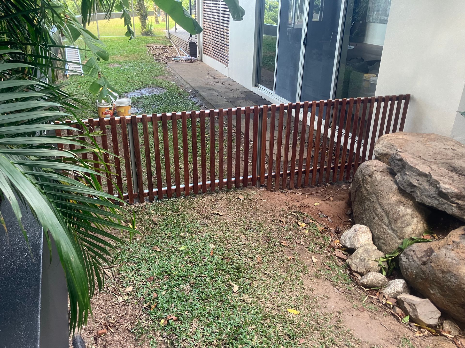 Small fenced yard with grass, a wooden gate, and large rocks beside a house.