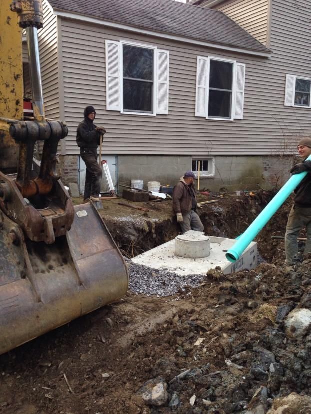 installing a septic system. A backhoe is in foreground; men are digging and connecting pipes near a house.