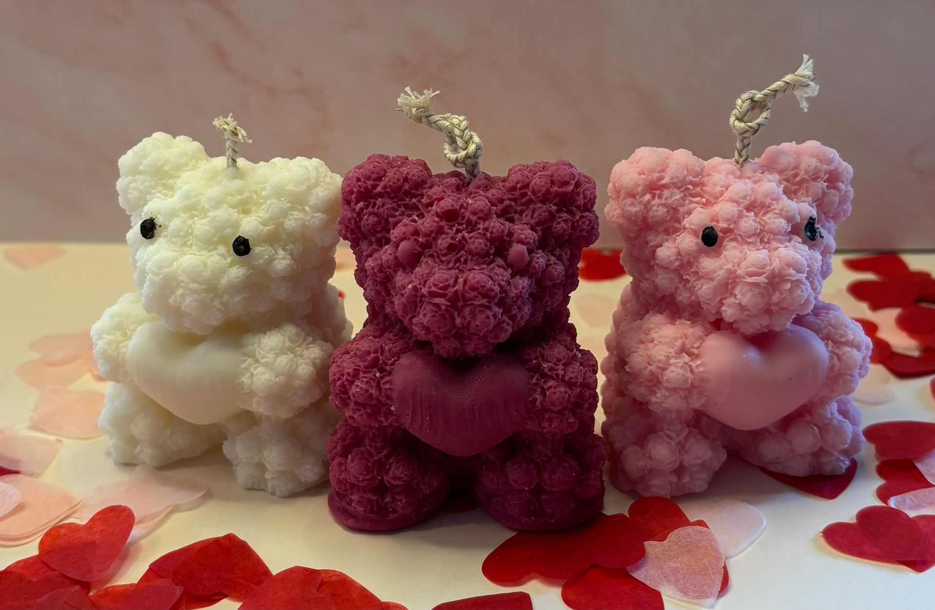 Drie teddybeerfiguurtjes kaarsen in crème, bordeauxrood en roze met hartjes op een tafel.