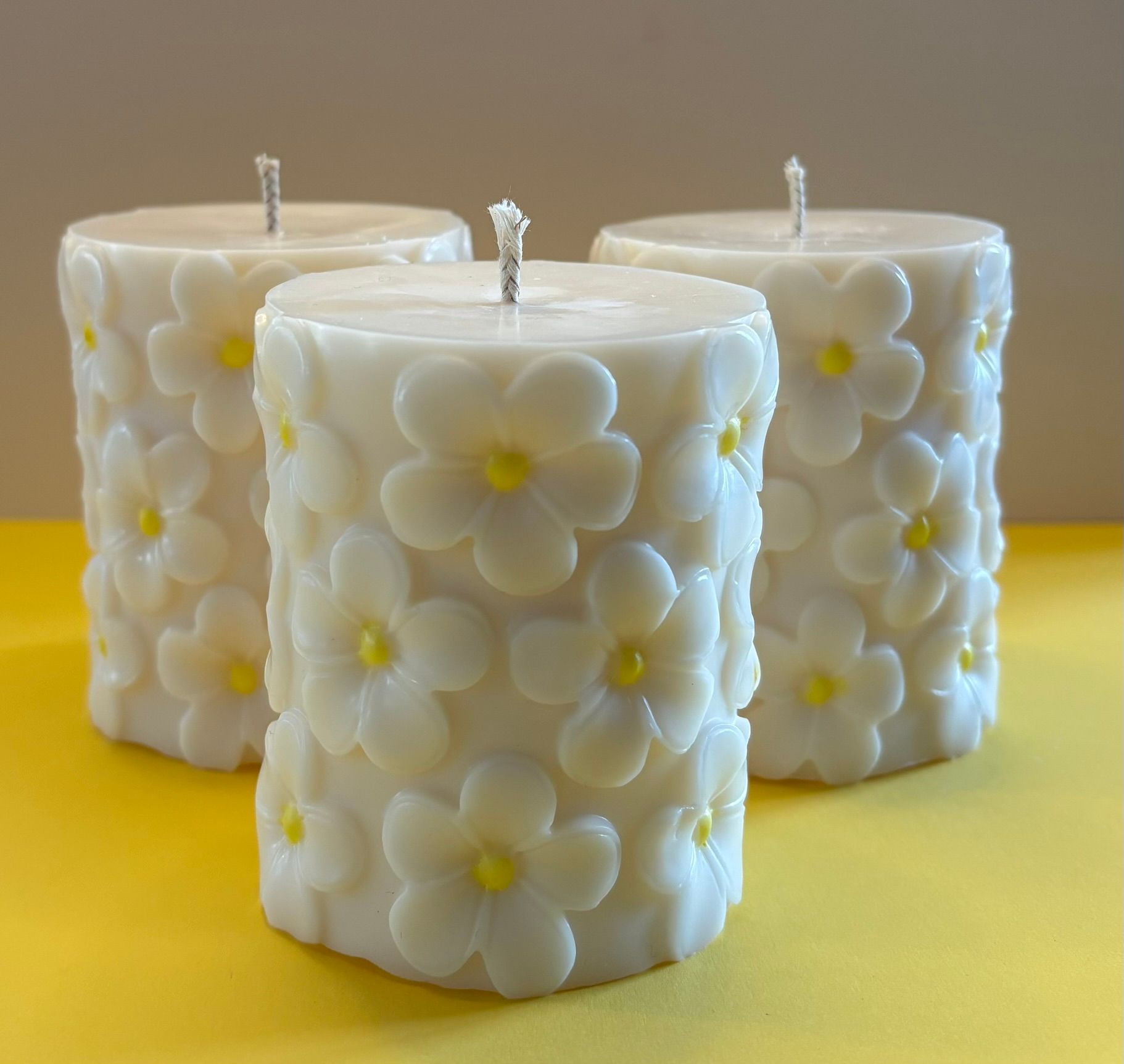 Drie witte kaarsen met bloemmotief op een gele ondergrond, met één kaars in het midden vooraan.