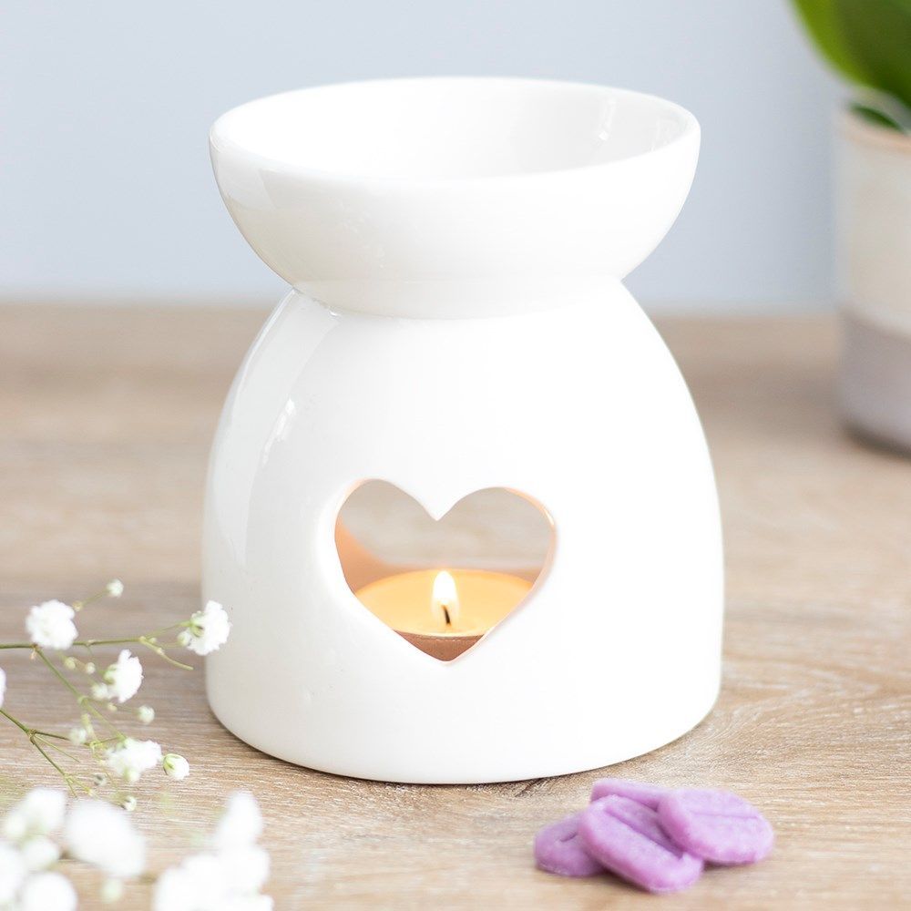 Een witte keramische waxbrander met een hartvormige uitsparing, een brandend theelichtje erin en kleine paarse waxblokjes op een tafeltje.