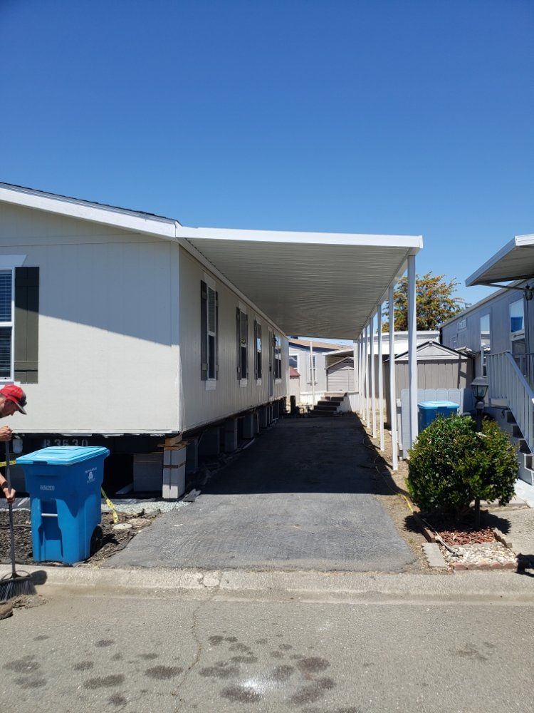 A man is standing in front of a mobile home with a covered driveway.