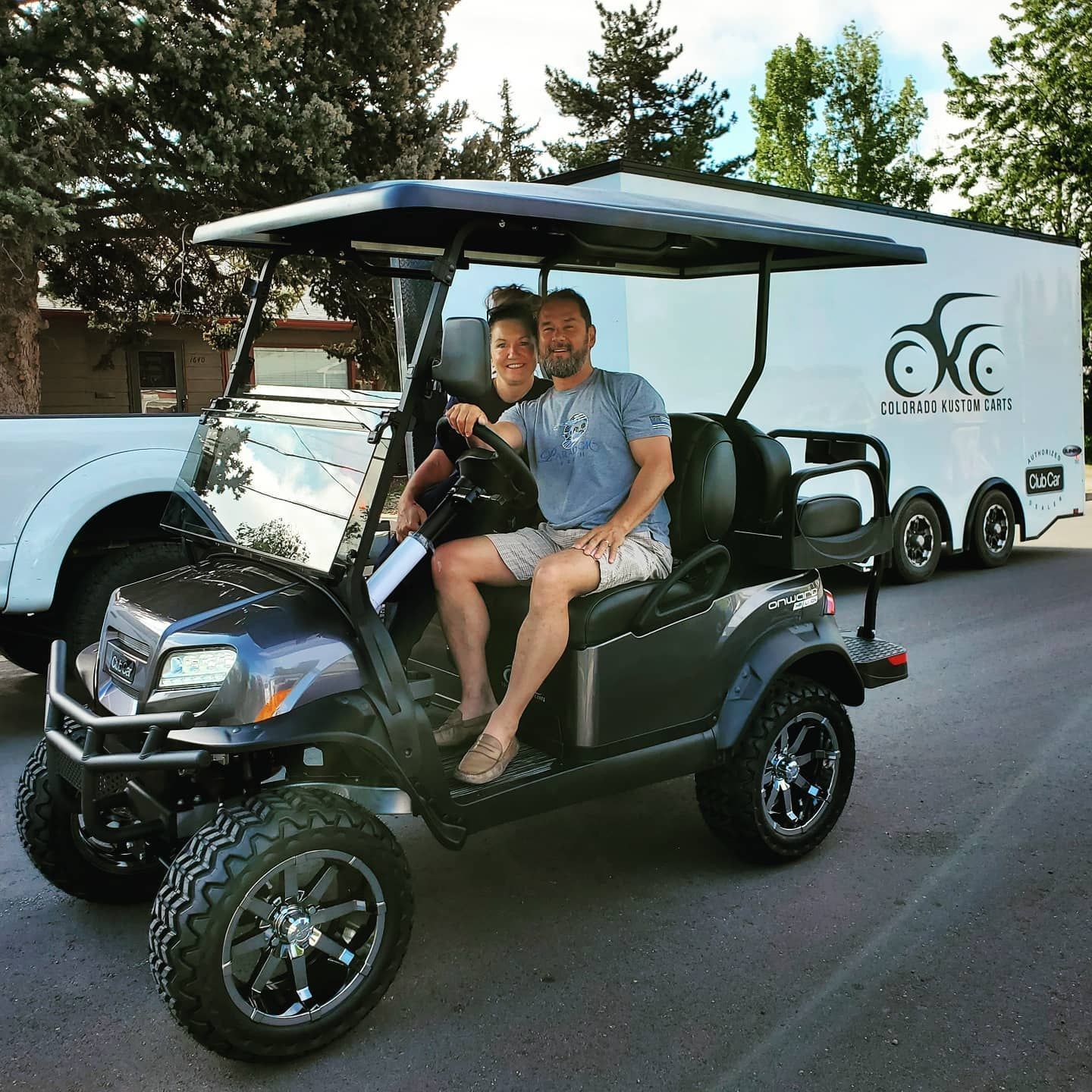 Two people sitting in a black golf cart, smiling on a street with a trailer in the background