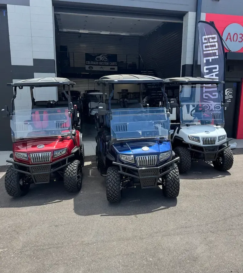 Three golf carts parked outside a shop, including red, blue, and silver vehicles.