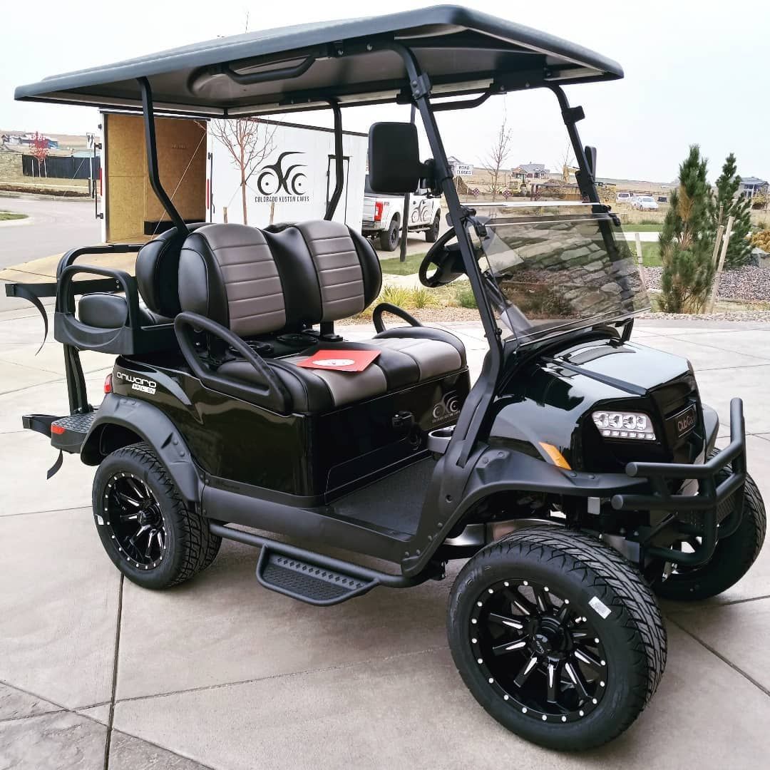 Black golf cart with canopy and lifted tires parked on a concrete lot near a building and shrubs