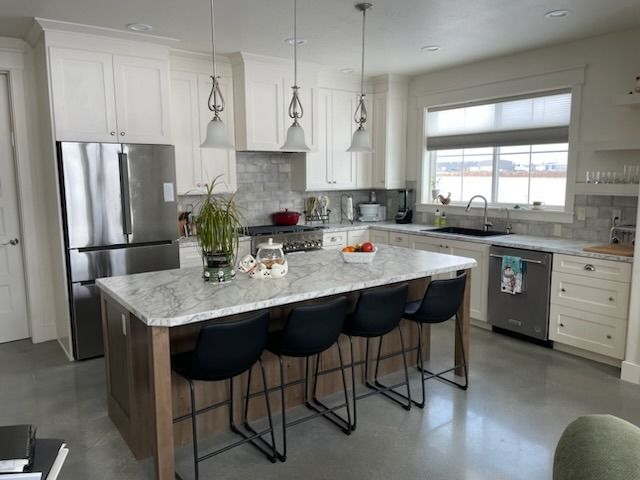 Modern white kitchen with marble island, black stools, stainless fridge, and pendant lights