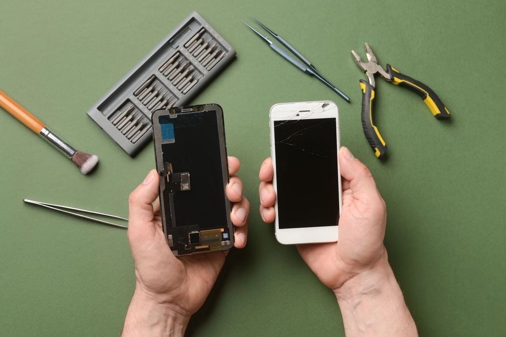 Mani che reggono un telefono nero danneggiato e un telefono bianco, con strumenti di riparazione su un banco da lavoro verde.