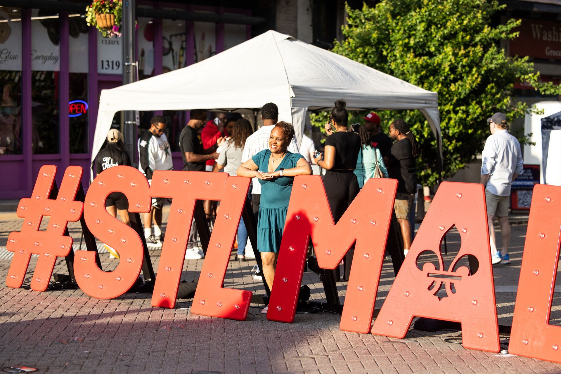 People gathered near large orange “#STLMA” letters and a white canopy tent outdoors