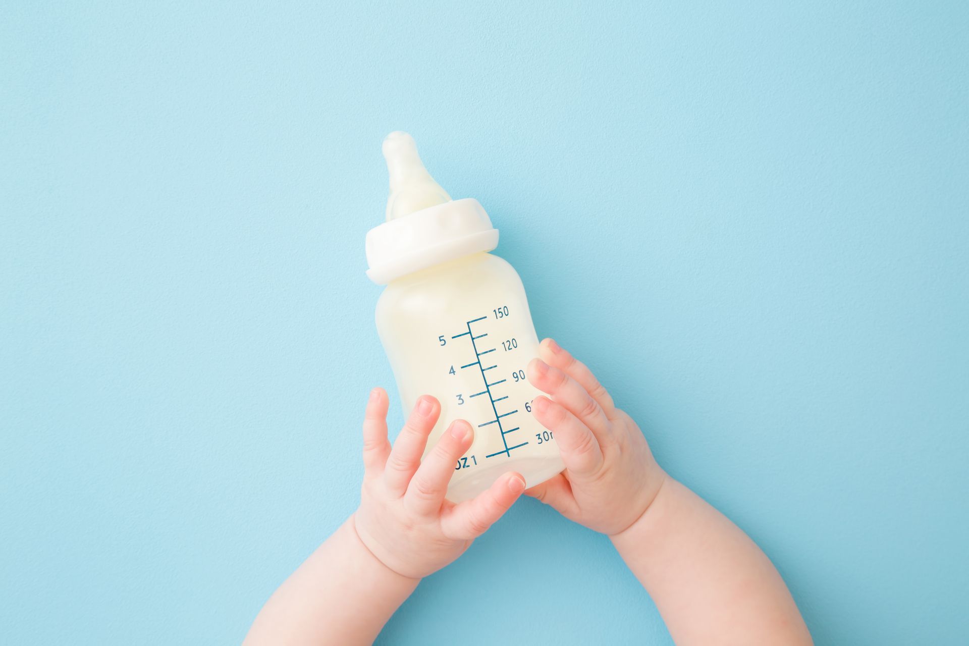 Baby hands holding a clear bottle of milk on a light blue background