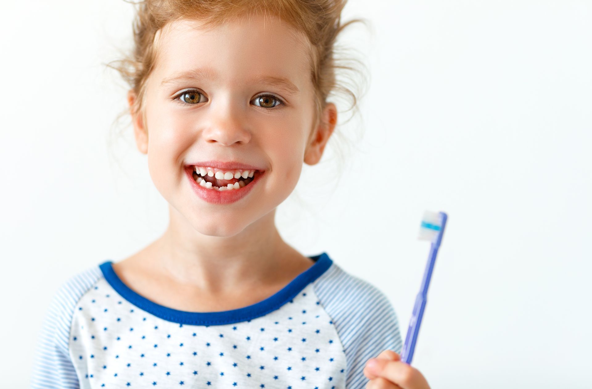 Une jeune fille sourit, sans une dent de devant, tout en tenant une brosse à dents, sur fond blanc.