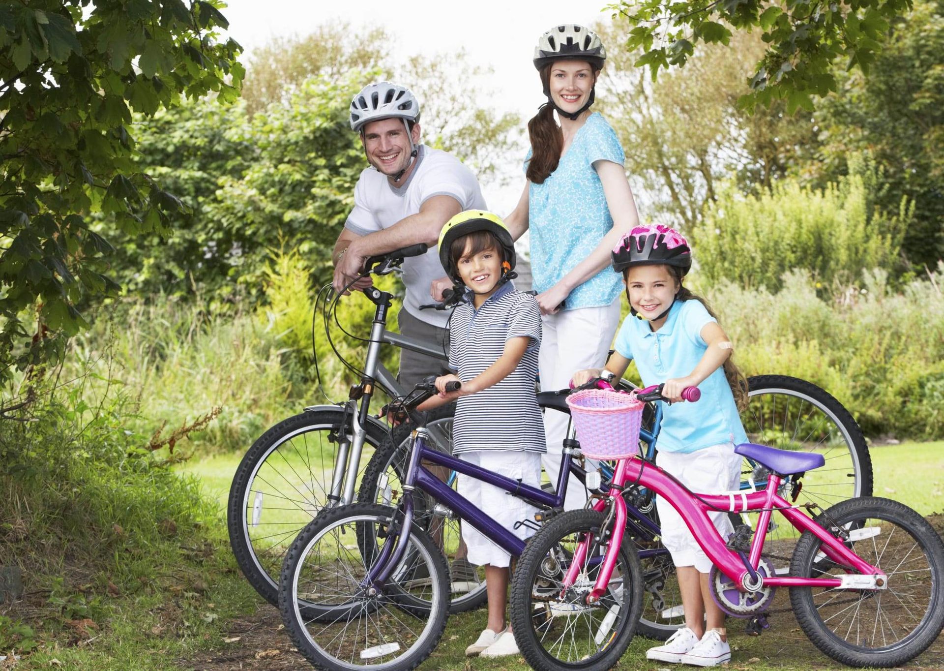Four cyclists with helmets and two bikes pose on a grassy trail, smiling in a sunny park.