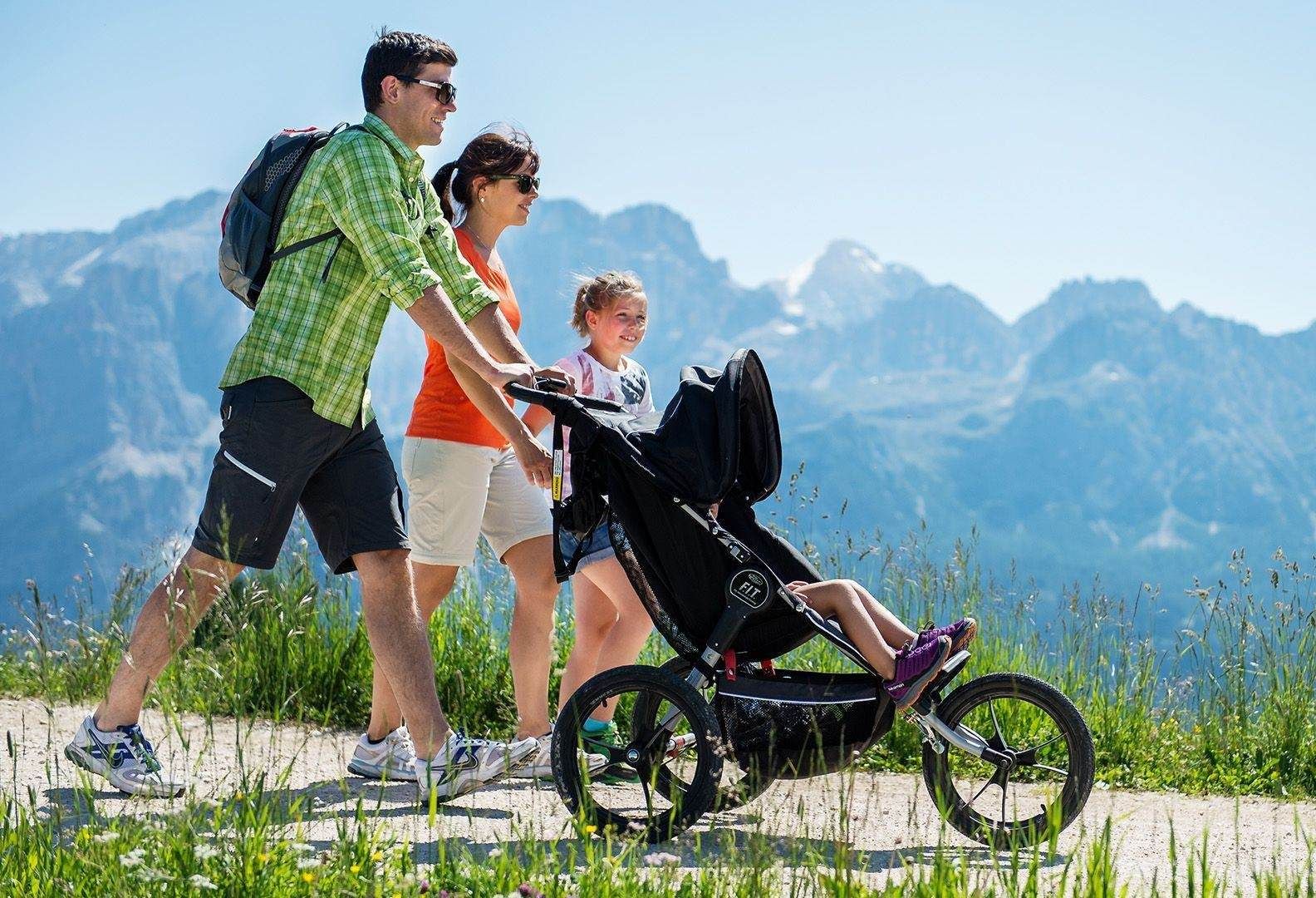 Three hikers walking with a stroller on a mountain trail under a clear blue sky
