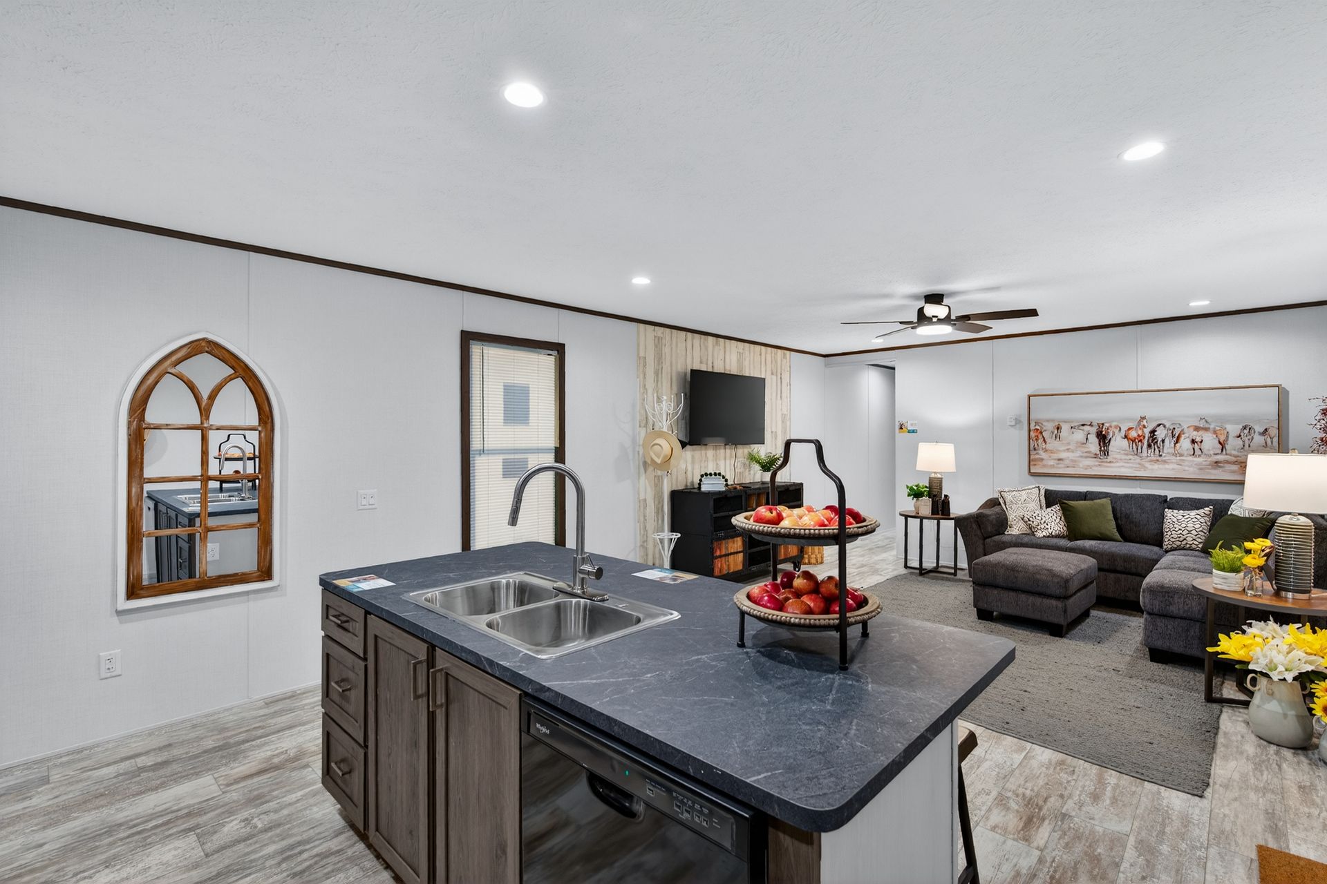 Open-concept kitchen and living room with gray decor, island sink, sofa, wall TV, and floral centerpiece.