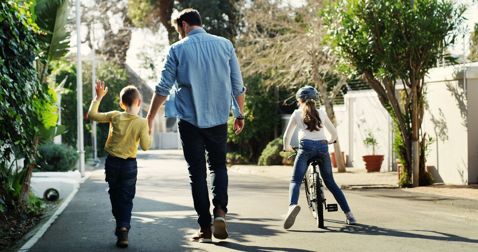 Three people walk down a sunny suburban street, two children holding hands with an adult.