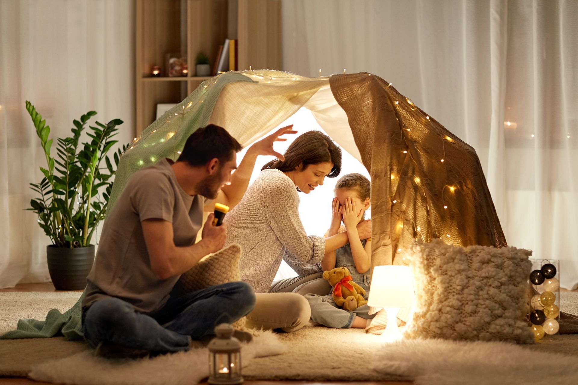 Family in a blanket fort with warm lights, reading together on a cozy living room floor.