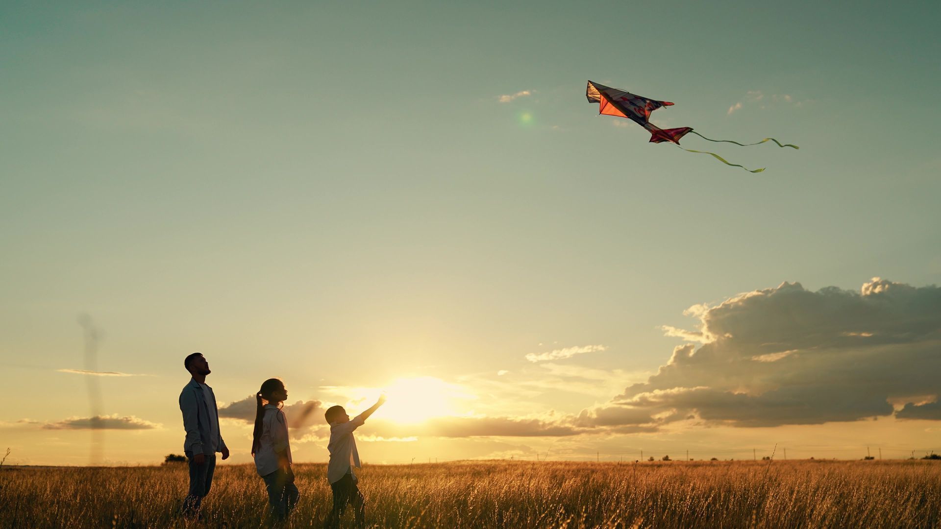 Three people fly a kite in a sunlit field at sunset.