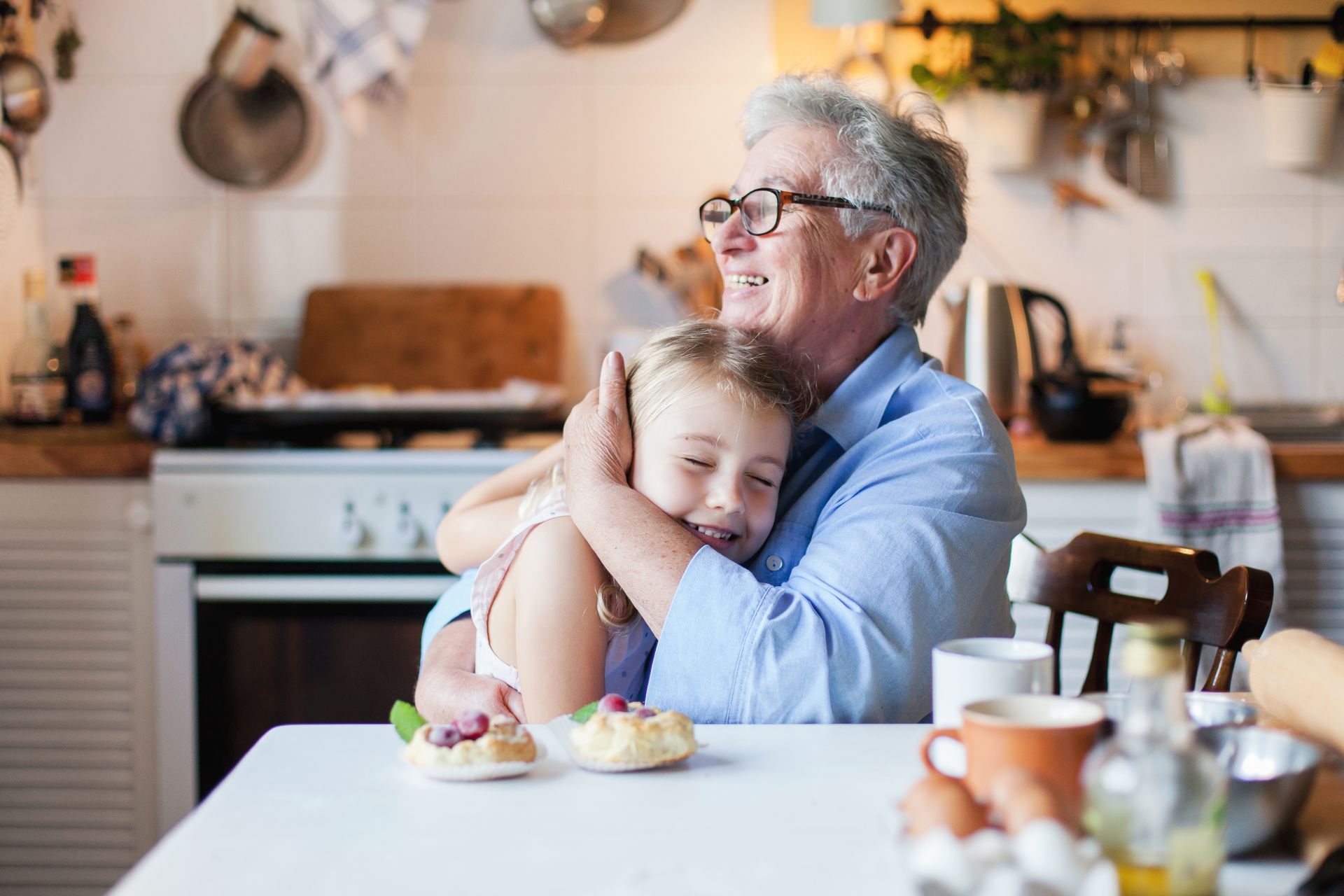 Older adult hugging a child at a kitchen table with cupcakes and coffee in a warm home kitchen