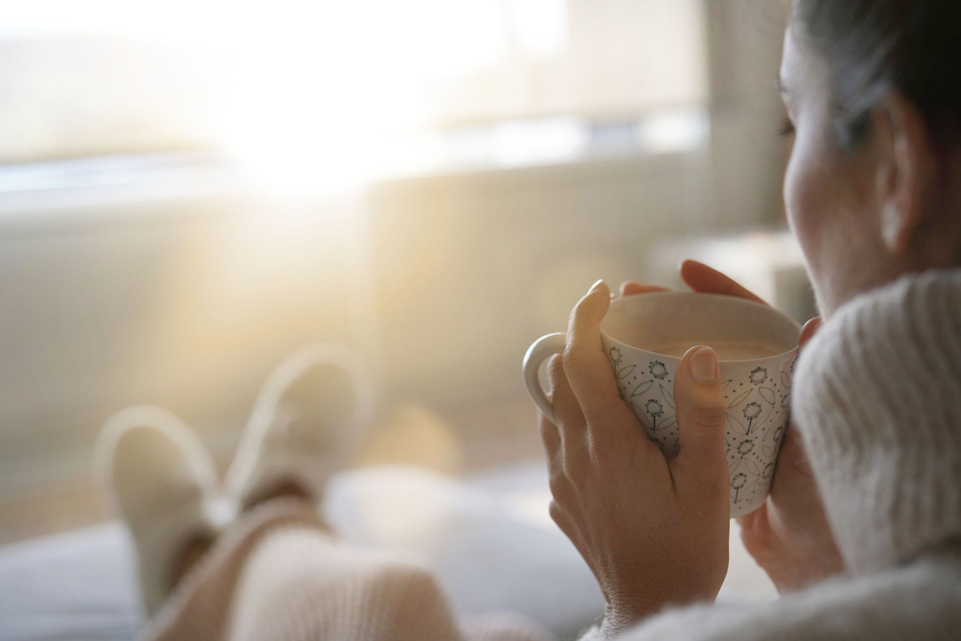 Person holding a steaming mug in a cozy, sunlit room with feet up on a sofa