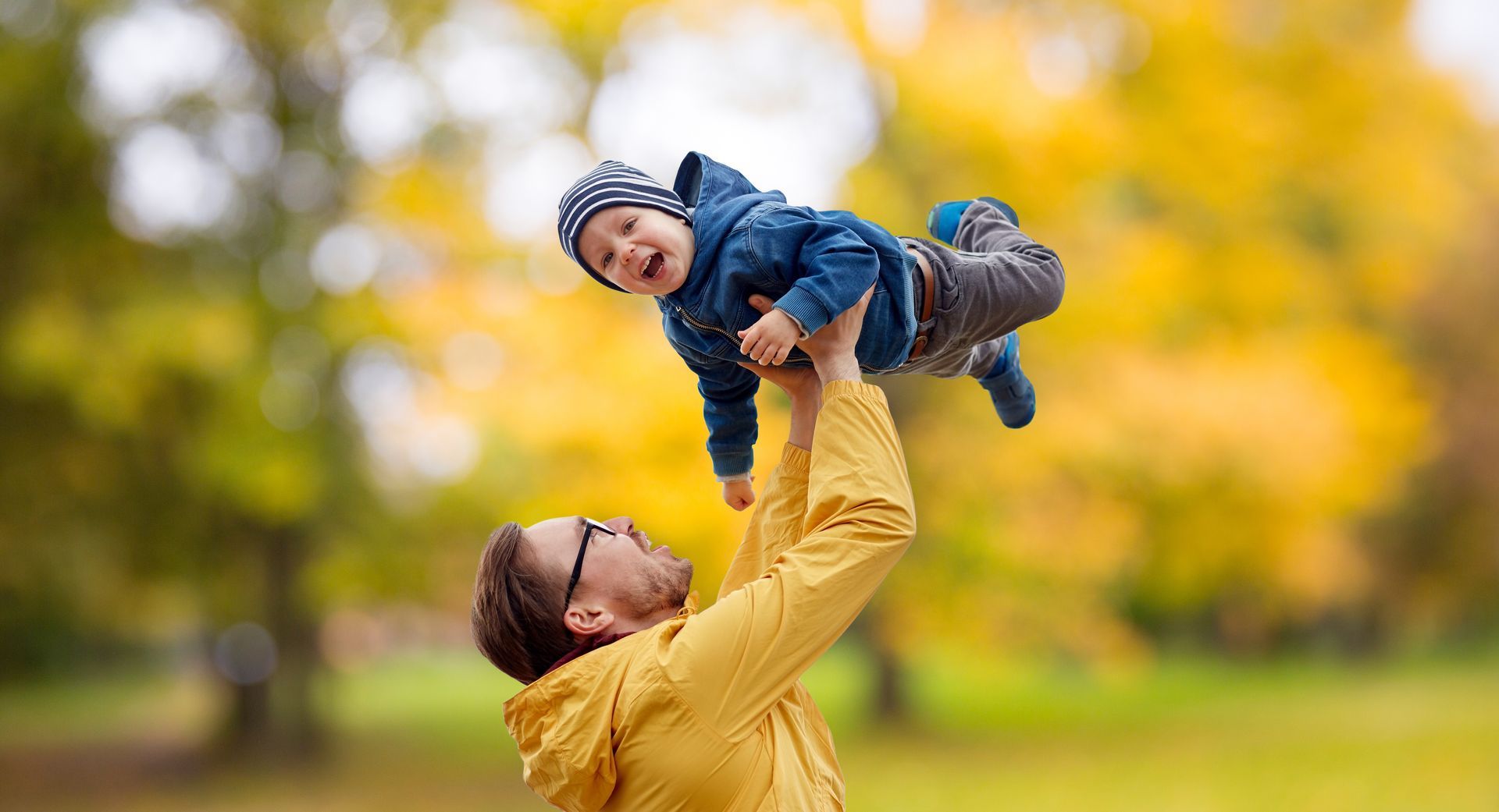 Adult lifting a smiling child in the air outdoors with golden autumn trees in the background
