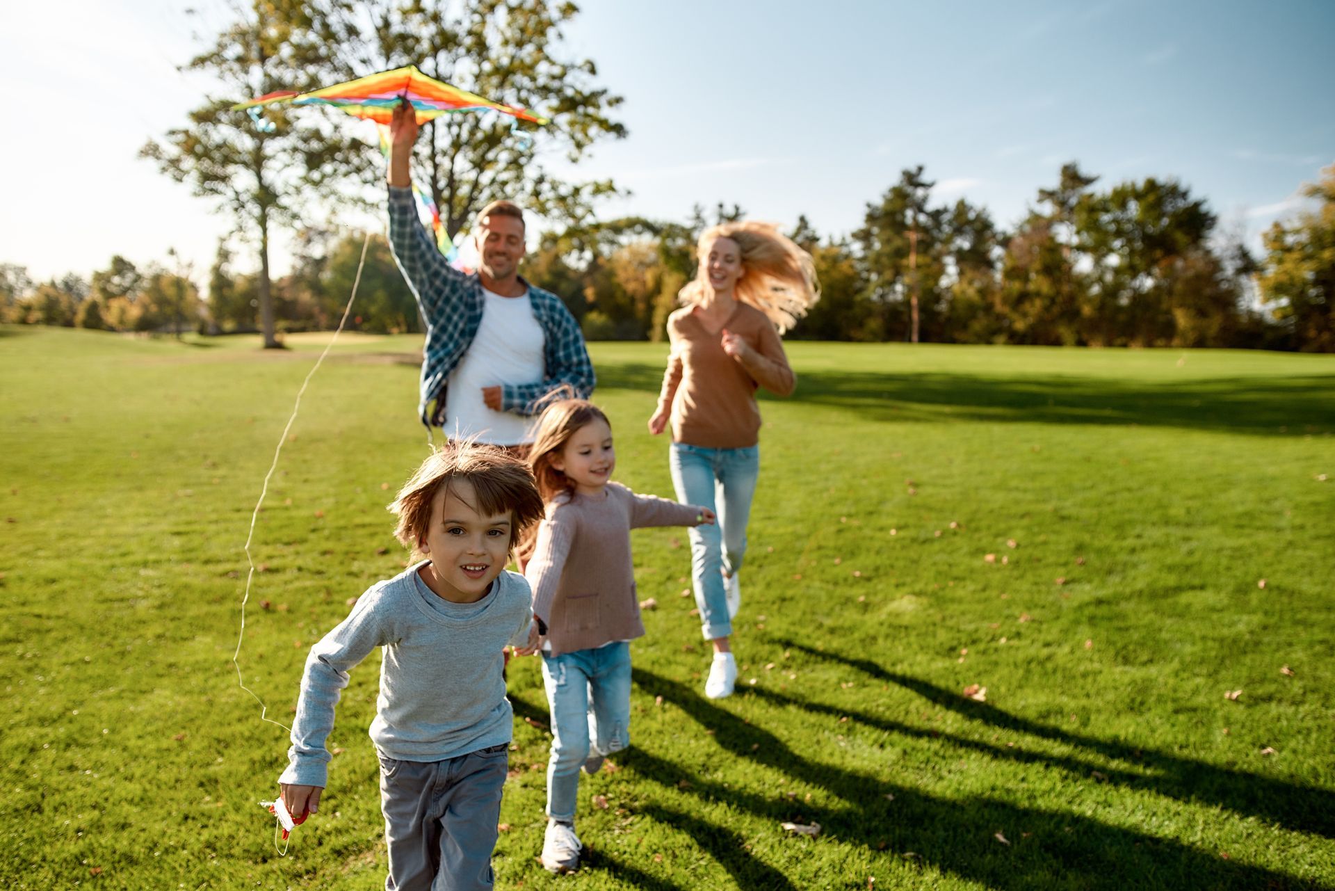 Children flying a colorful kite in a sunny park, with one adult walking behind them