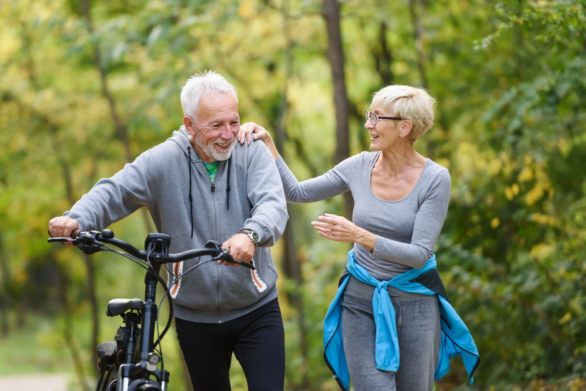 Two people smiling beside a bicycle in a park, one wearing a gray hoodie and the other a blue sweater tied at the waist