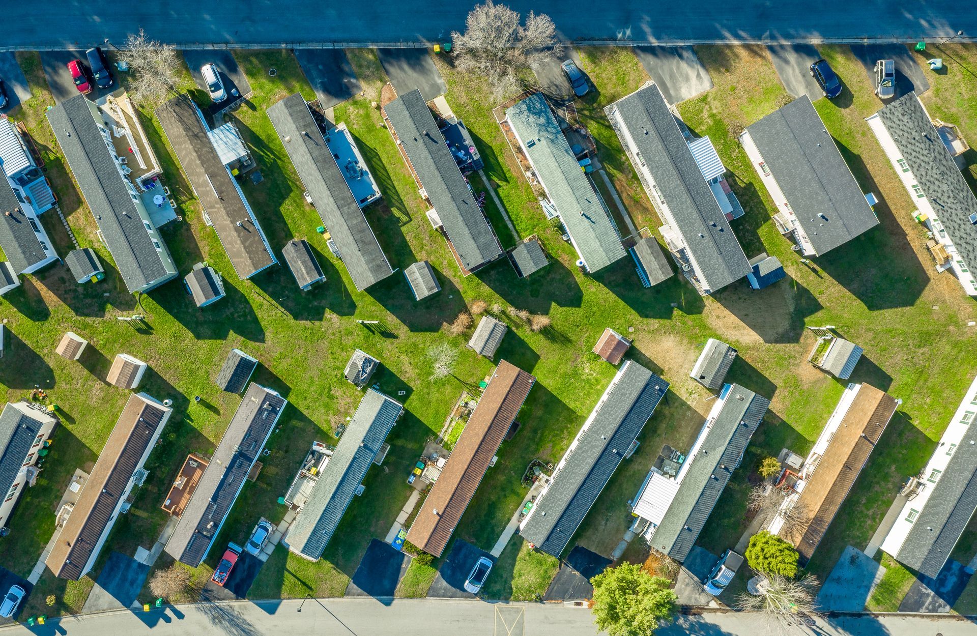 Aerial view of a mobile home park with rows of beige trailers, green lawns, and trees near water.