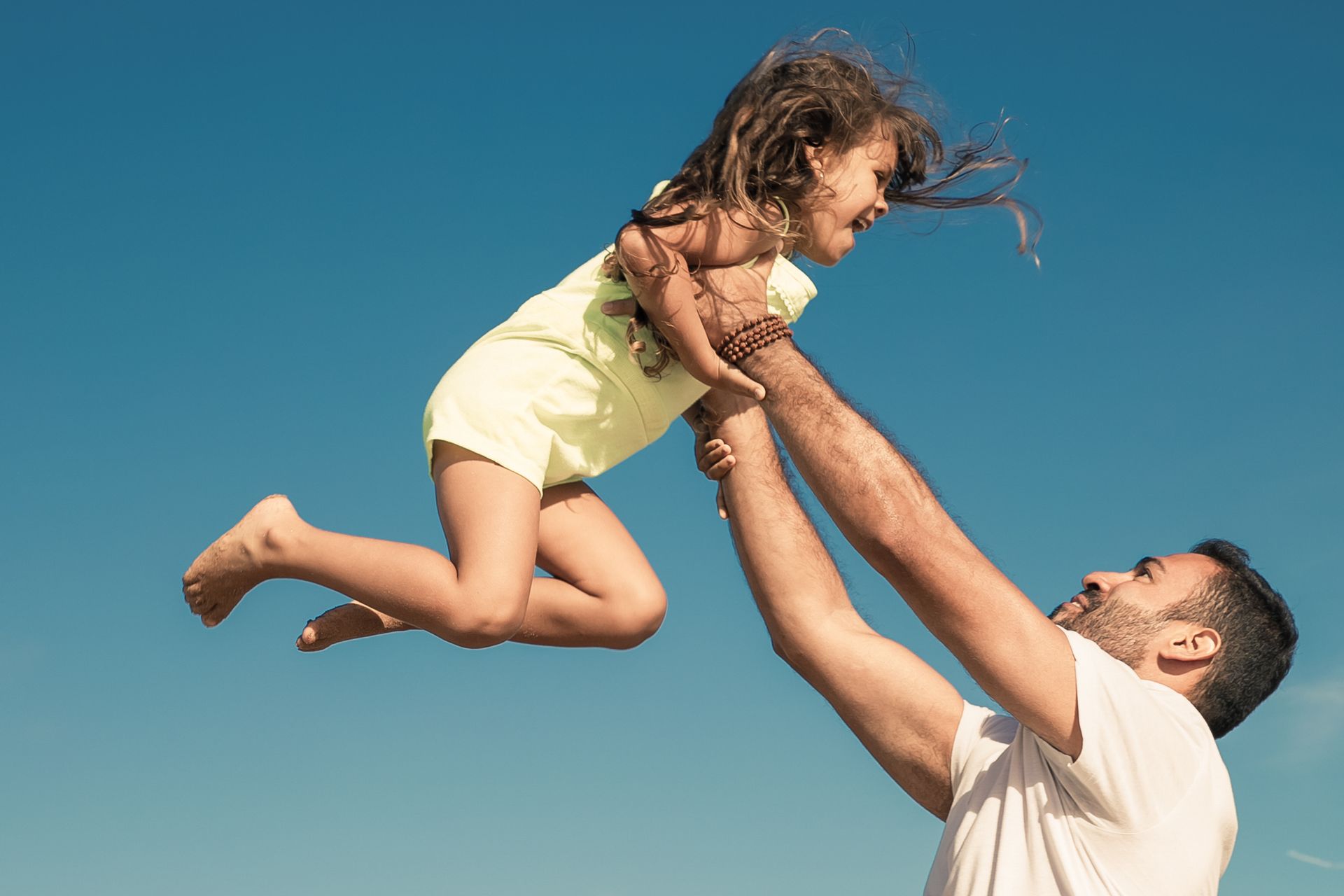 Adult lifting a smiling child into the air against a clear blue sky