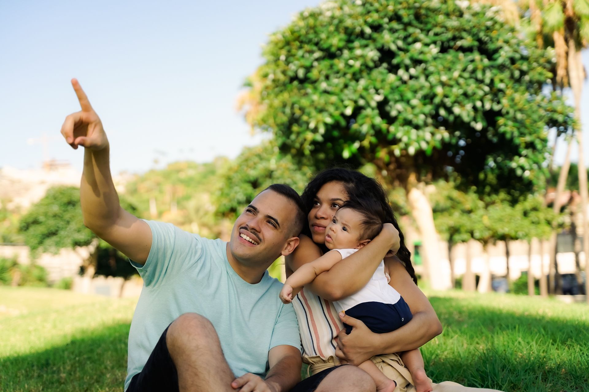 Smiling couple on grass, one pointing ahead, with a green park tree in the background