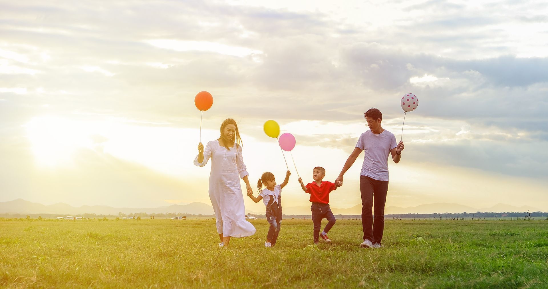 Family walking with colorful balloons across a sunny grassy field at sunset