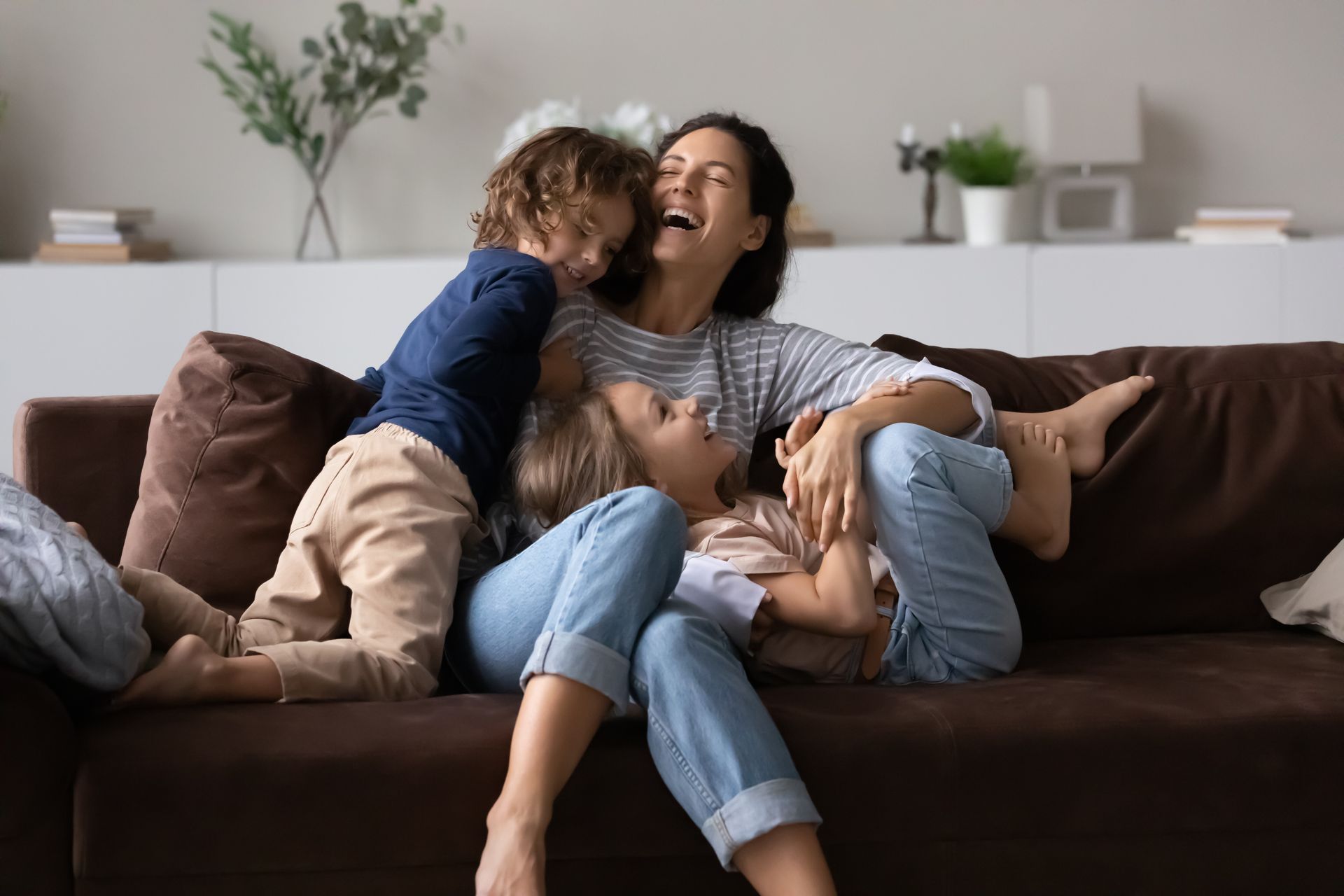 A person laughing on a couch with two playful children in a bright living room.