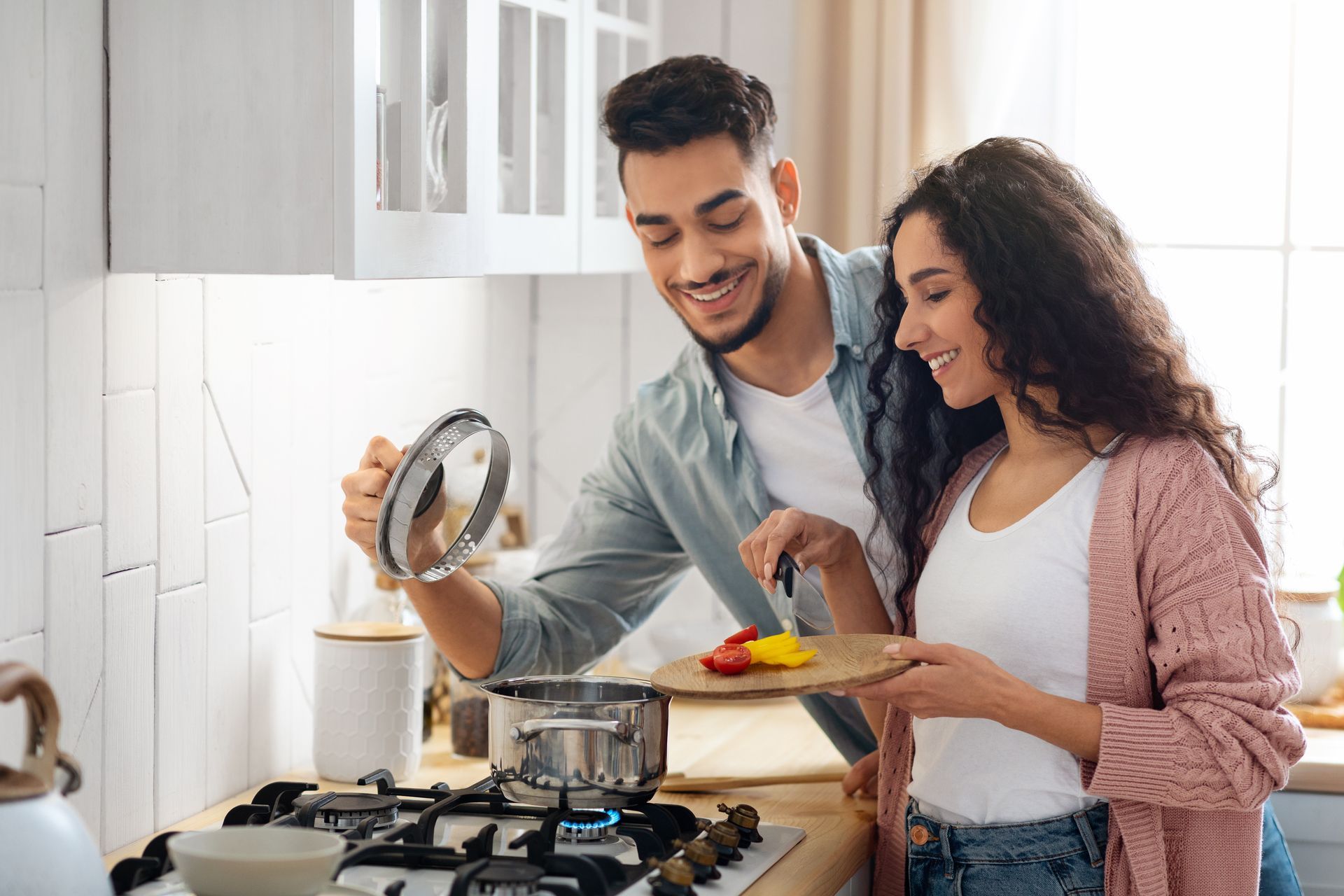 Two people cooking together in a bright kitchen, smiling while holding a pot lid and spatula.