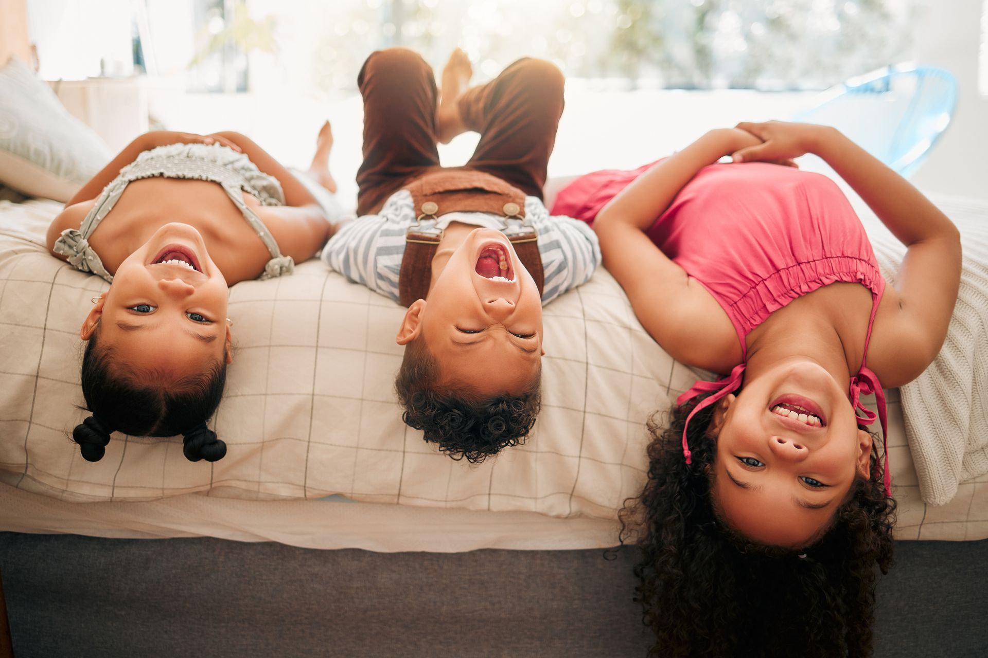 Three children lying upside down on a bed, smiling and looking at the camera.