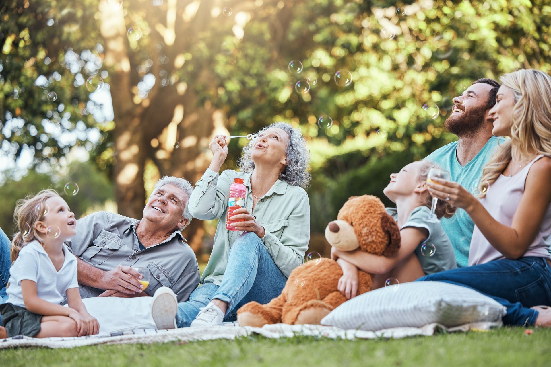 Friends relaxing on a sunny lawn with a teddy bear and drinks, enjoying a picnic outdoors