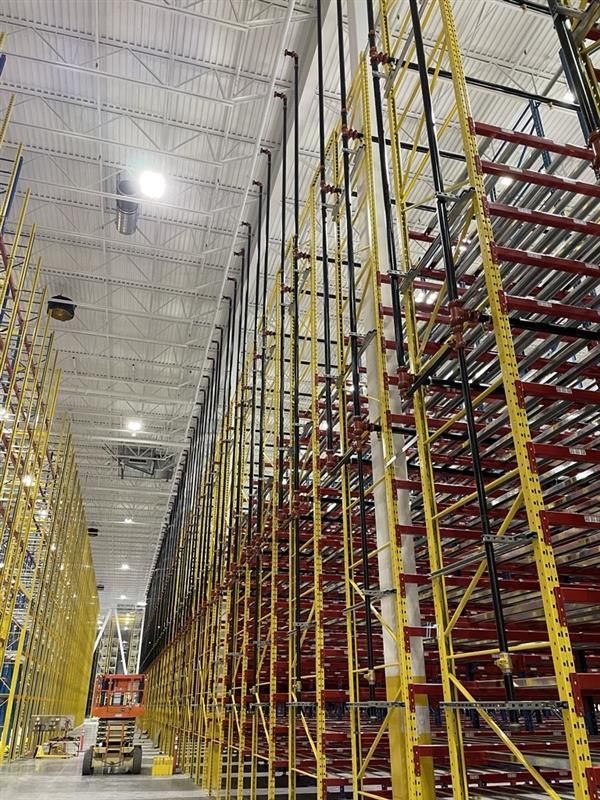 Aisle in a warehouse with tall yellow shelving, red racks, and an orange forklift under bright lights.