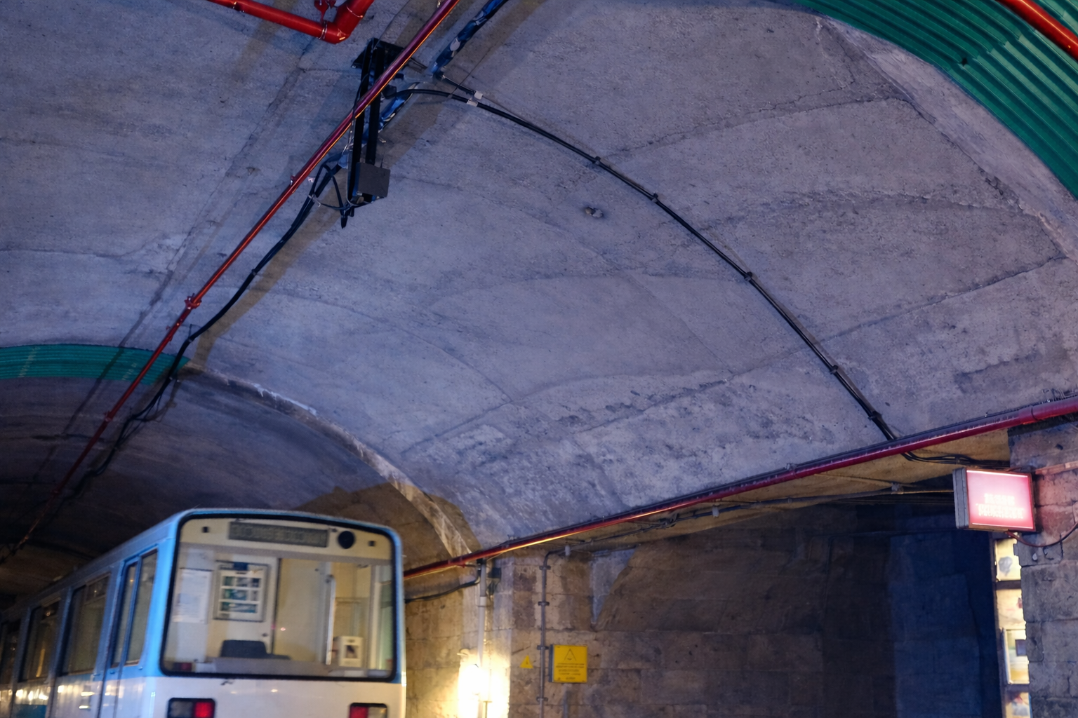 Subway train entering a concrete tunnel with overhead cables and a lit station sign