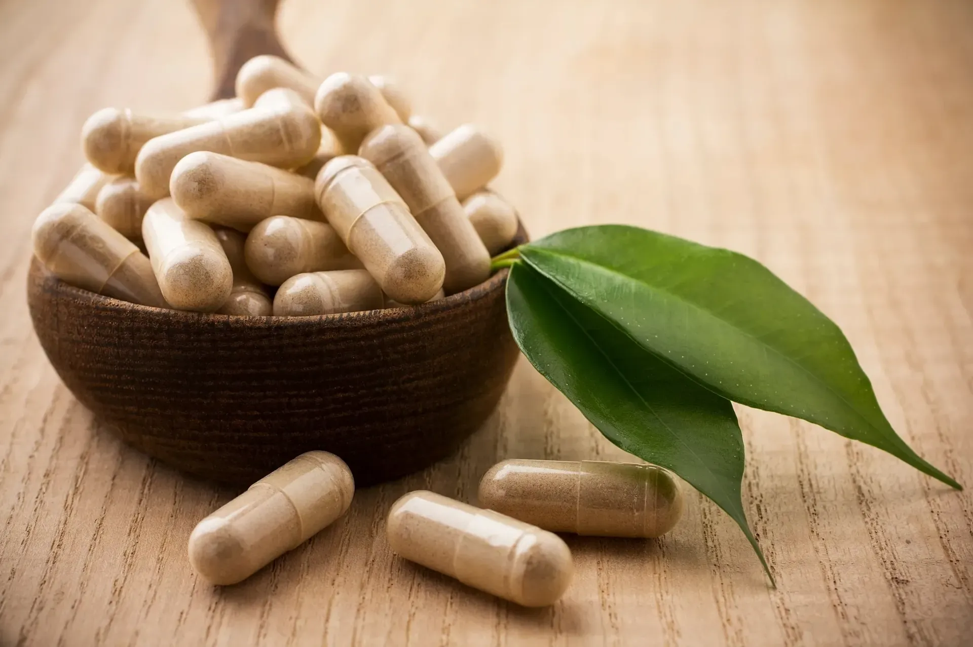 Herbal capsules in a bowl beside green leaves on a wooden table