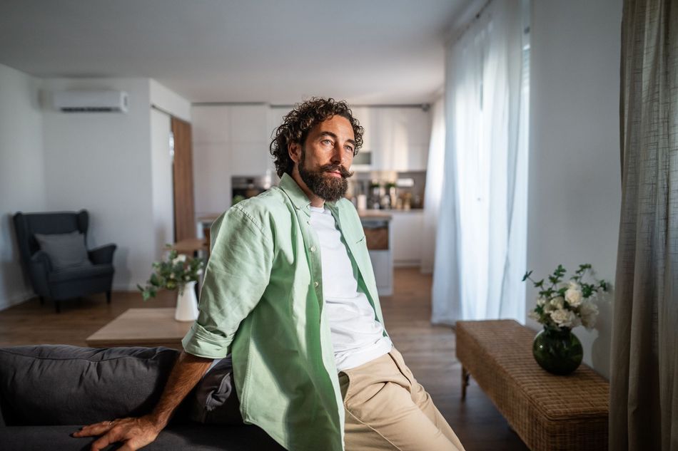 Man sitting on a sofa in a bright living room with a green shirt and curtained windows nearby
