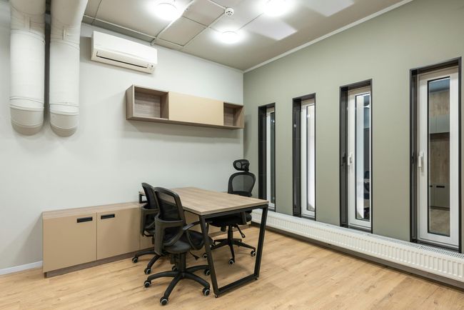 Small office with desk, chairs, wall shelves, filing cabinet, and tall windows under bright lights