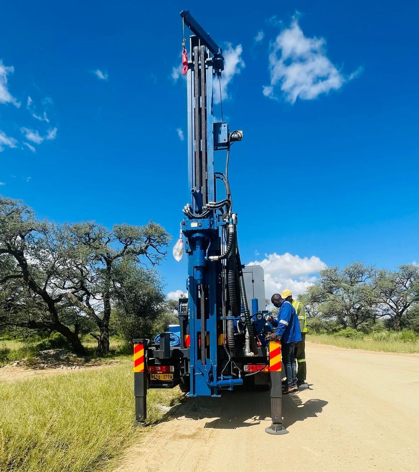 Blue drilling rig on a dirt road, under a blue sky. A person in blue work clothes is nearby.