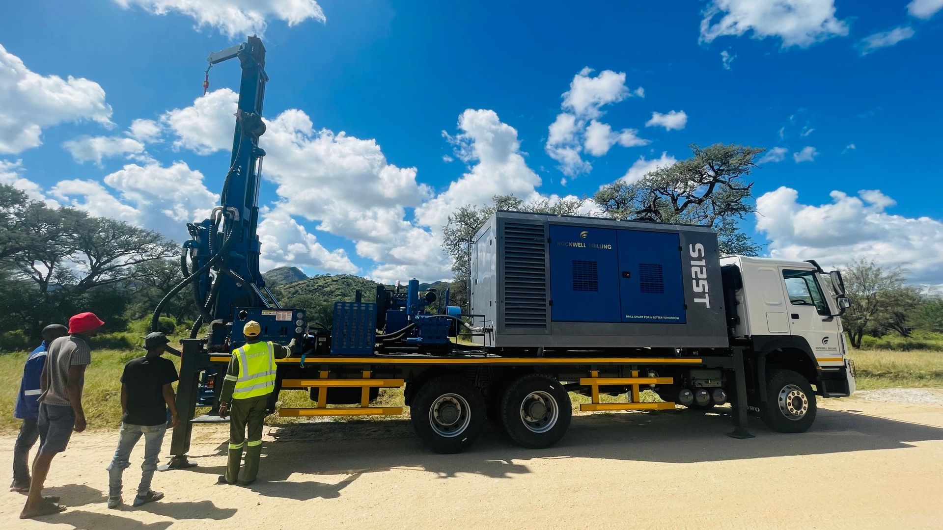 Truck-mounted drilling rig with workers, under a blue sky.