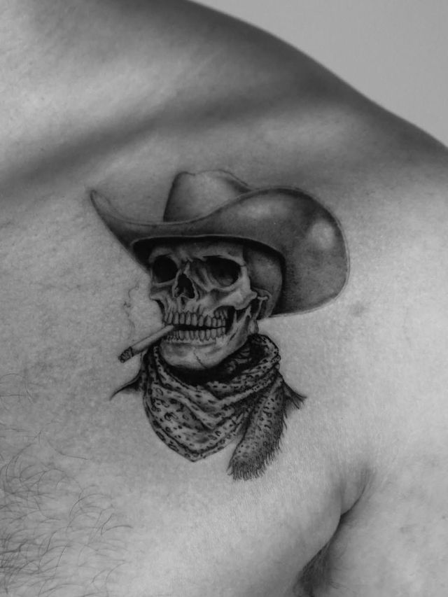 A grayscale tattoo on a shoulder depicting a skull wearing a cowboy hat and a bandana while smoking a cigarette.